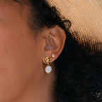 Close-up of an ear with Knot Pearl Drop Earrings in 18K Gold, featuring a minimalist design with a round white pearl and subtle knot detail, person wearing a hat.
