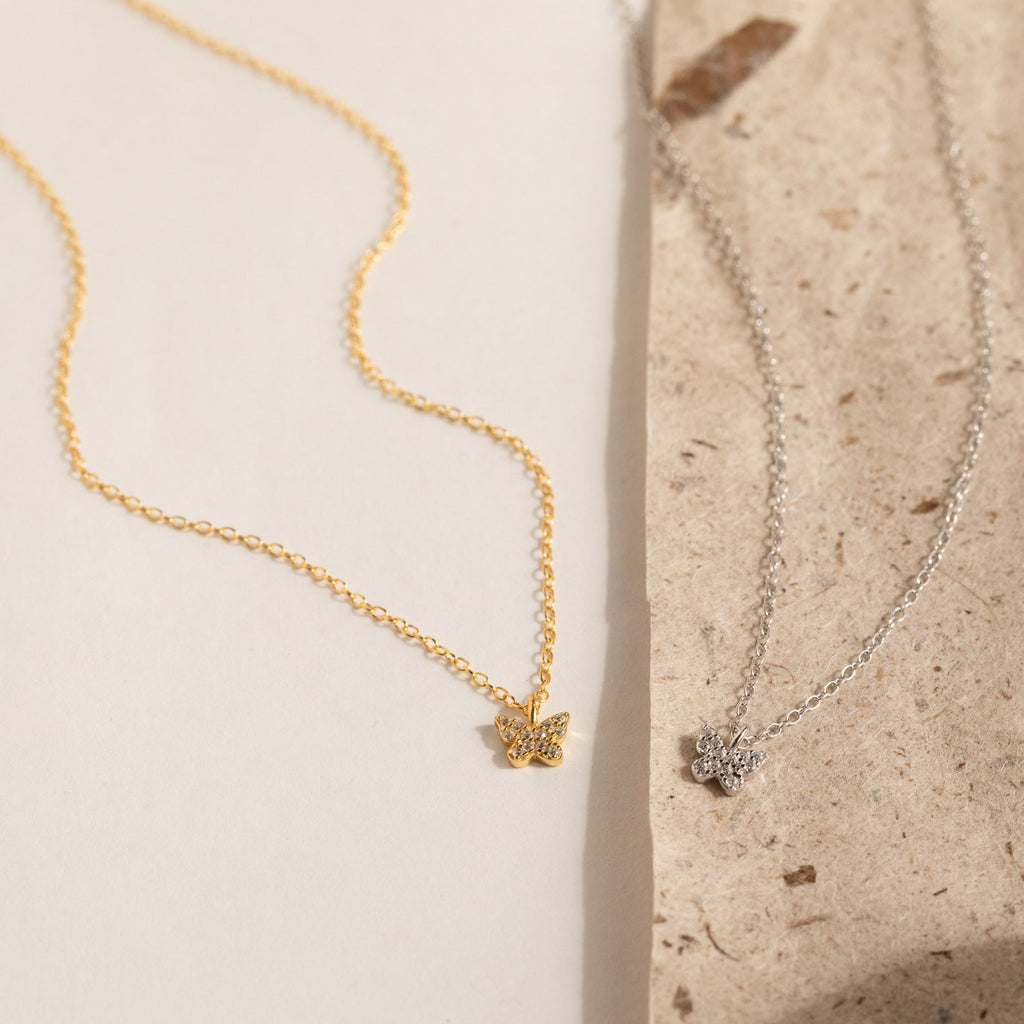 The Mandy Pave Butterfly Necklace, available in gold or silver and embellished with sparkling diamonds, is displayed on a textured cream and beige background.