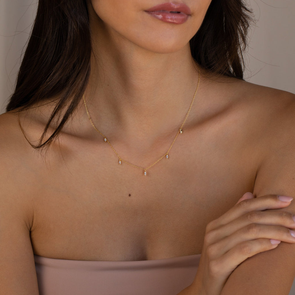 A woman wears the Marquise Station Necklace, featuring marquise and teardrop diamond charms on a delicate chain, with a strapless nude top and her arms gently crossed.