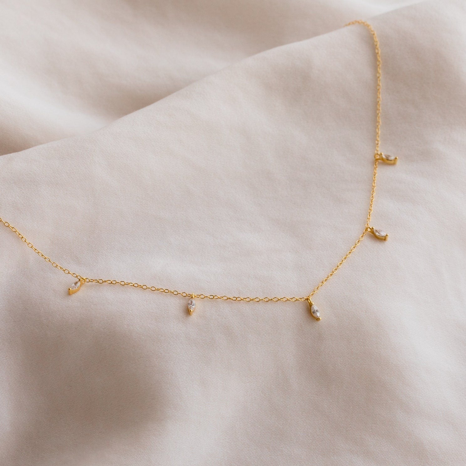 The Marquise Station Necklace is a delicate gold chain adorned with small marquise diamond charms, elegantly displayed on soft beige fabric.