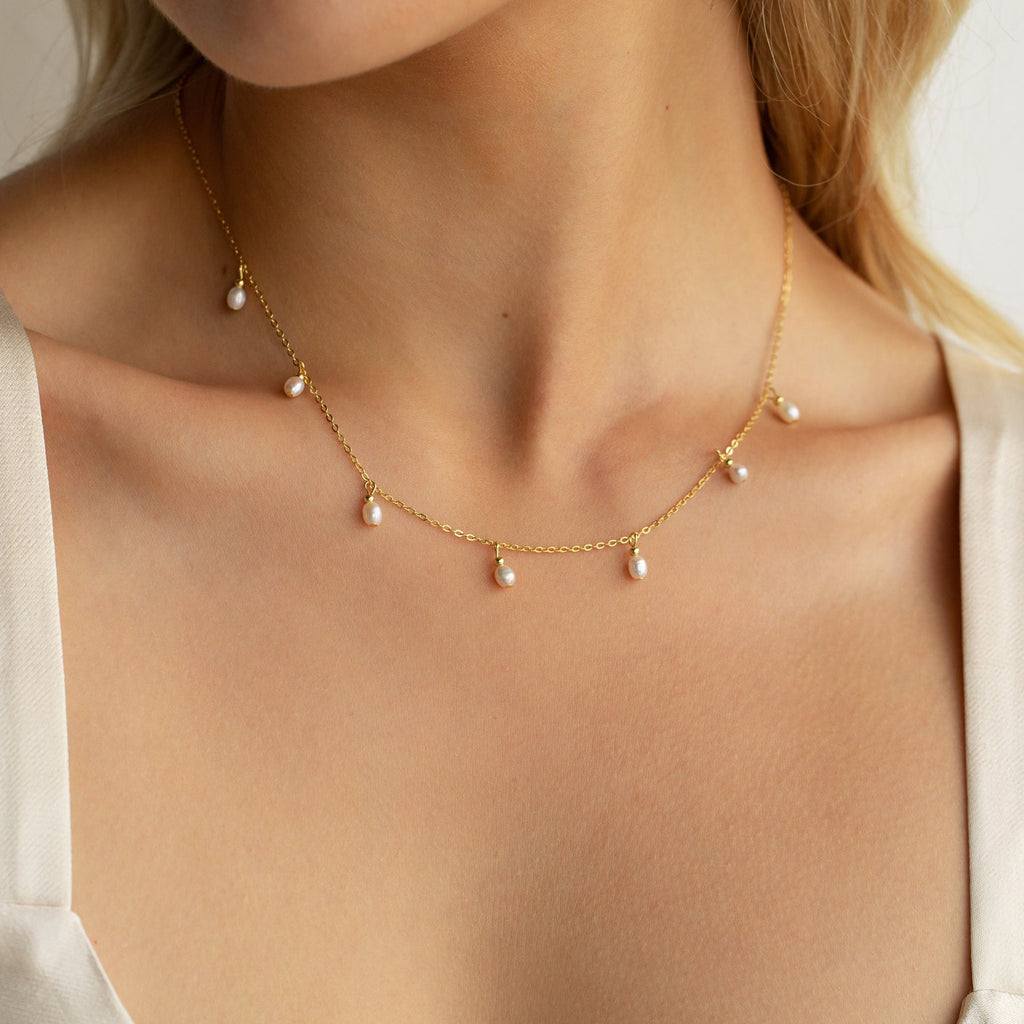 A close up of our Pearl Station Necklace in 18K Gold finish on our models neck, hanging just along the collarbone - featuring seven small 5mm dangling Pearl Bead Charms spaced out along a dainty cable chain.