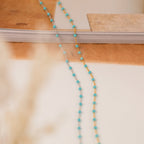 The Turquoise Station Necklace features delicate blue and gold beads, shown on a white and brown surface near stacked books—a charming accessory for any style.