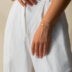 Wearing white pants, a person rests their hand on their hip, highlighting three Essential Bracelet Chains—delicate gold designs with polished finishes.