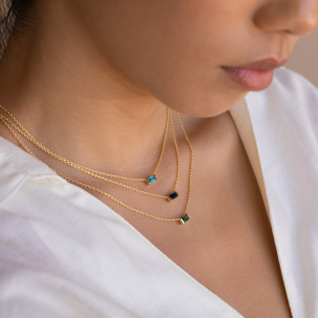 A close-up of model wearing three layered Emery Birthstone Necklaces in gold, each with a different rectangular gemstone in blue zircon, sapphire, and emerald