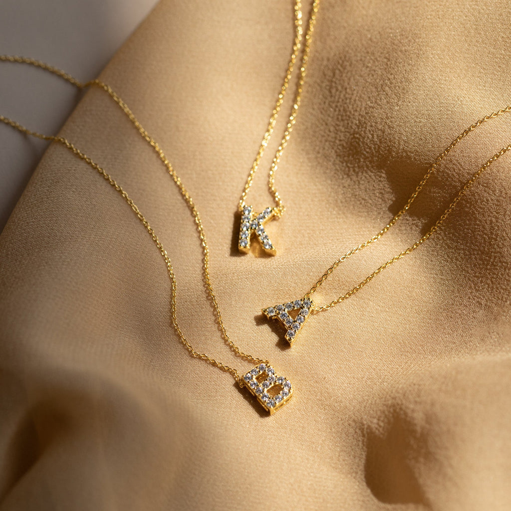 Three Floating Pave Initial Necklaces with diamond-encrusted K, A, and B letter pendants rest on beige fabric—an elegant display of personalized jewelry.