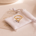 The Aphrodite Pave Heart Ring, featuring sparkling diamonds, sits on a white drawstring pouch decorated with an outline of two hands—an elegant anniversary gift.