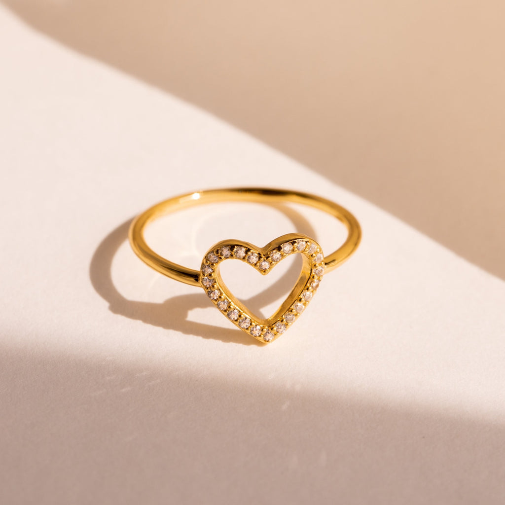 The Aphrodite Pave Heart Ring features an open heart outline paved with sparkling diamonds, shown on a cream-colored background—an ideal anniversary gift.