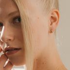 Close-up of a blonde woman wearing minimalist Amelia Onyx Duo Huggies—small gold hoops with onyx accents—gently touching her face.