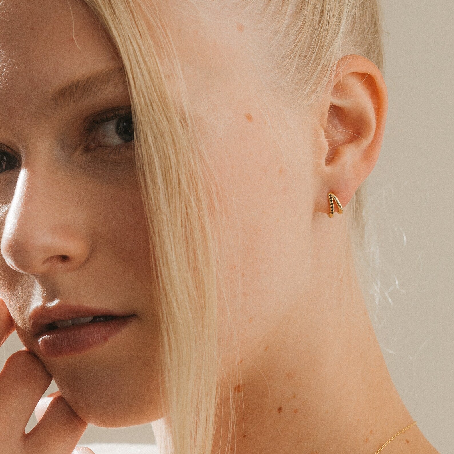 Close-up of a blonde woman wearing minimalist Amelia Onyx Duo Huggies—small gold hoops with onyx accents—gently touching her face.