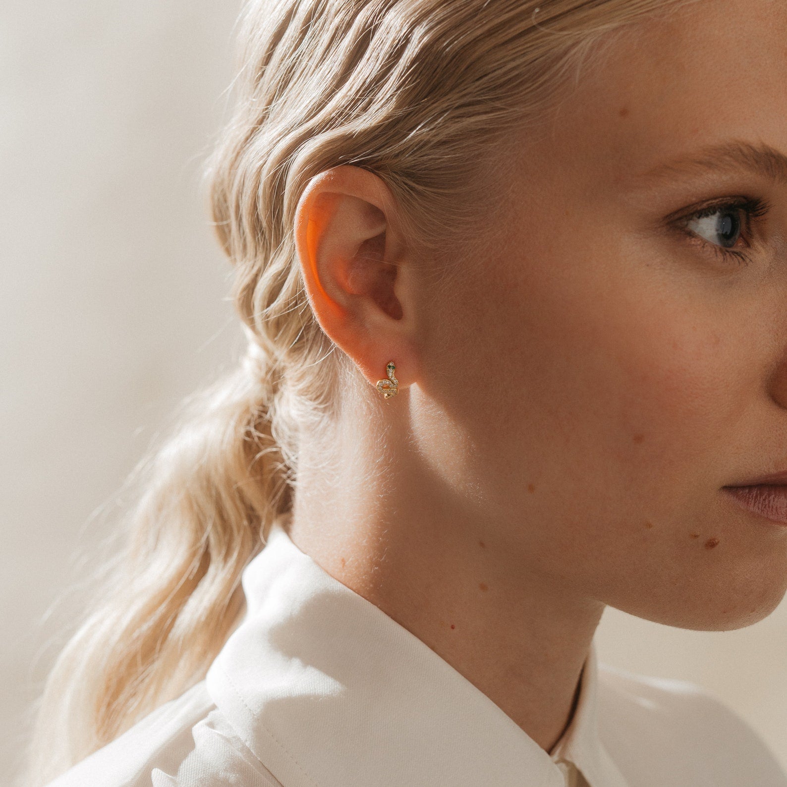 Close-up of a woman with blonde hair wearing trendy jewelry—the Cobra Snake Huggies gold stud earrings—and a crisp white collared shirt.