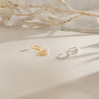 The Lila Pearl Huggies—dainty hoop earrings in gold and silver, each adorned with pearls—are displayed on a white surface with dried leaves, offering the perfect touch of modern jewelry elegance.