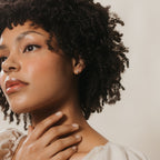 Close-up of a woman with curly hair and hand on her neck, thoughtfully gazing to the side, highlighting the Lila Pearl Huggies for a touch of modern jewelry elegance.