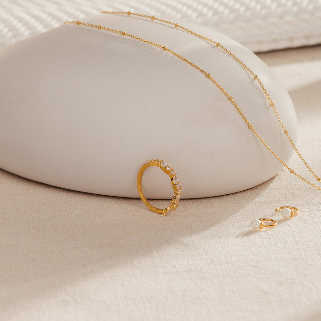 The Verity Diamond Ring, gold necklace, and pearl earrings are displayed on a smooth, beige surface.