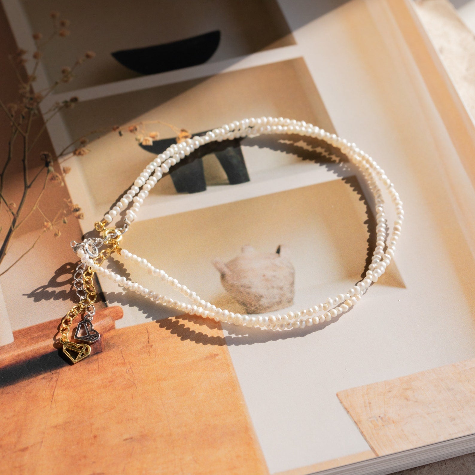 Delicate pearl anklet with tiny white beads, finished with a silver clasp and gold chain extender featuring small dangling heart charms, styled on a warm-toned background with soft sunlight.