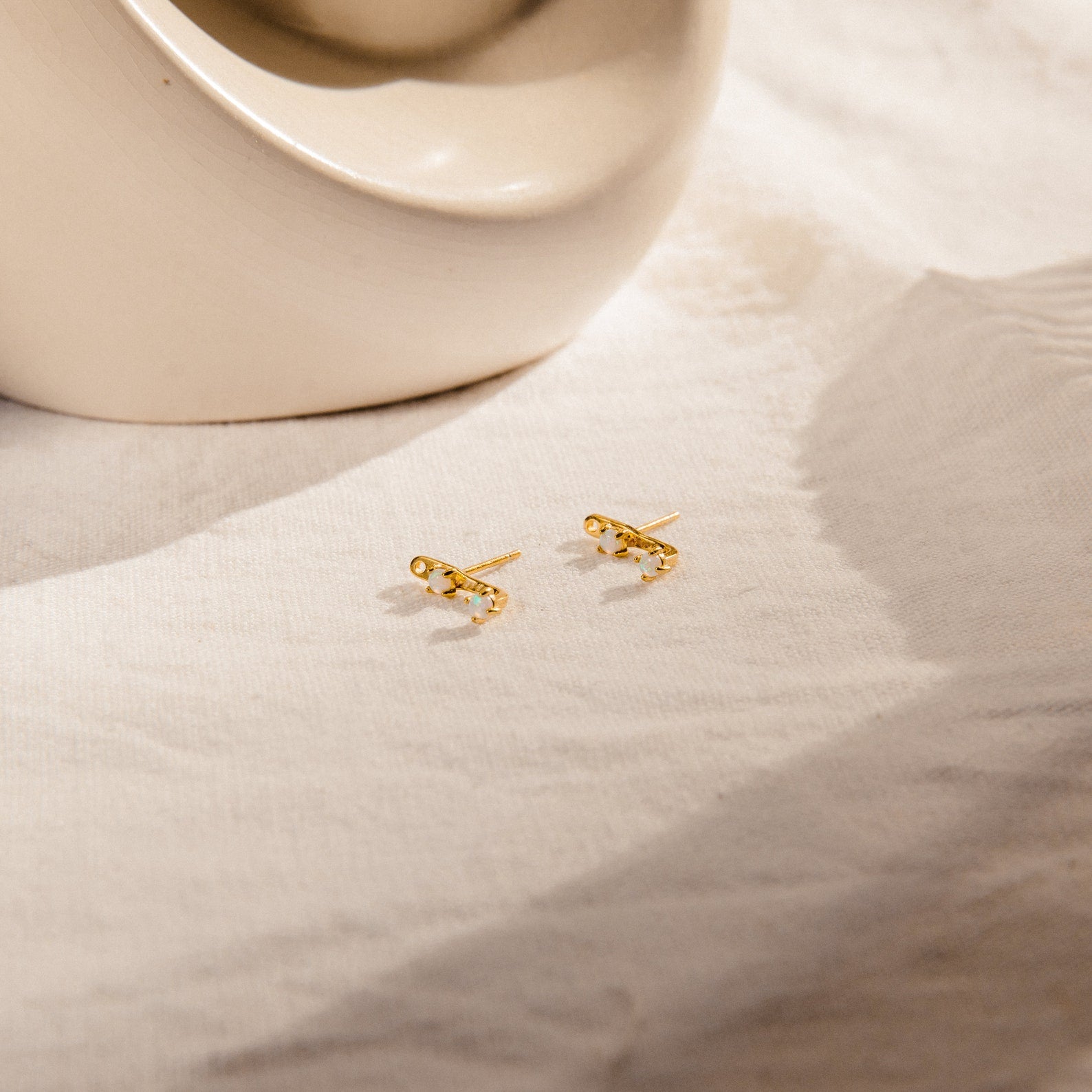 Opal Ear Jackets with small clear gemstones rest on cream-colored fabric beside a ceramic dish, featuring a removable earring post for versatile styling.