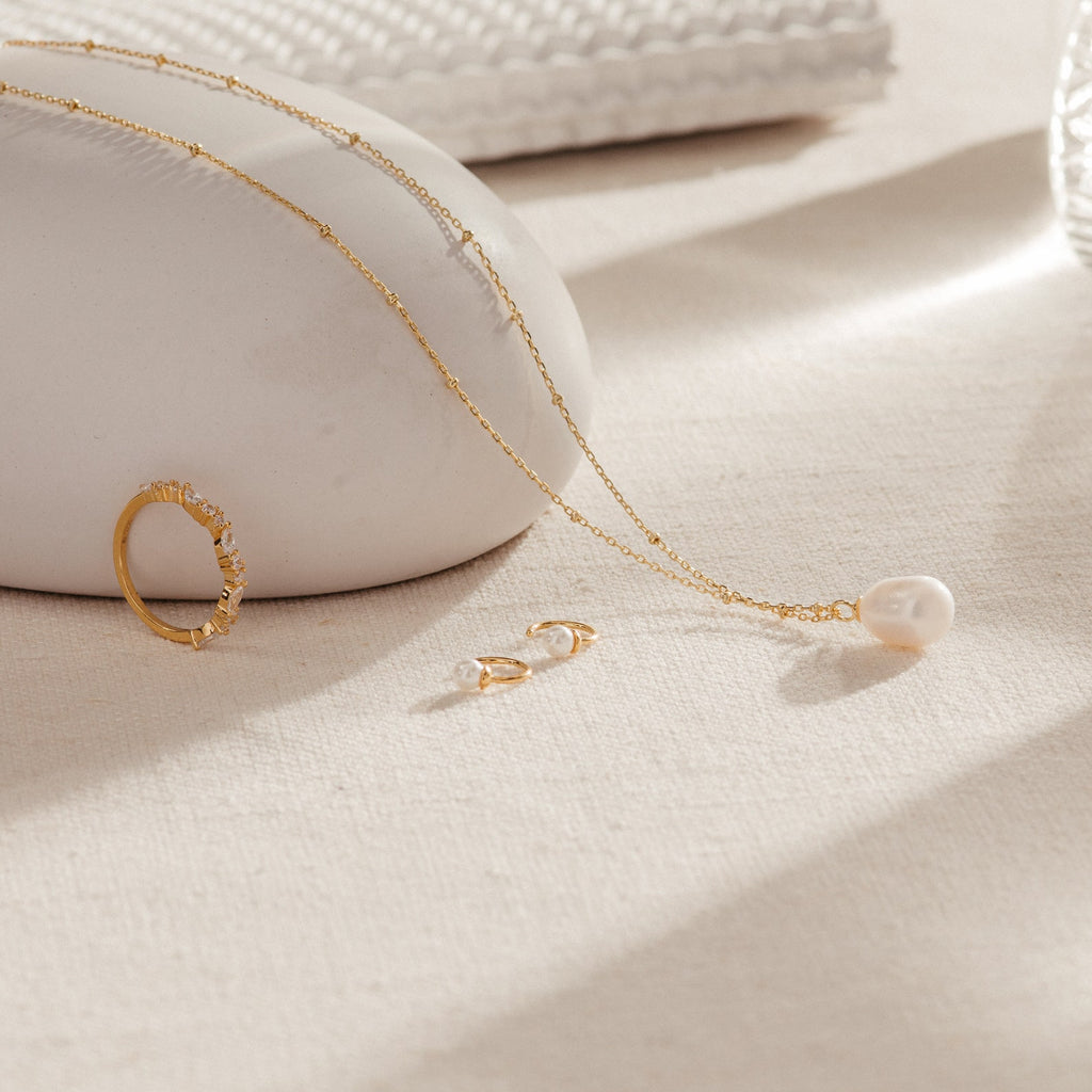 The Lila Pearl Huggies, a gold ring, and a pearl pendant necklace rest on a light beige surface with soft natural lighting—an elegant display of modern jewelry.