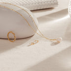 The Lila Pearl Huggies, a gold ring, and a pearl pendant necklace rest on a light beige surface with soft natural lighting—an elegant display of modern jewelry.