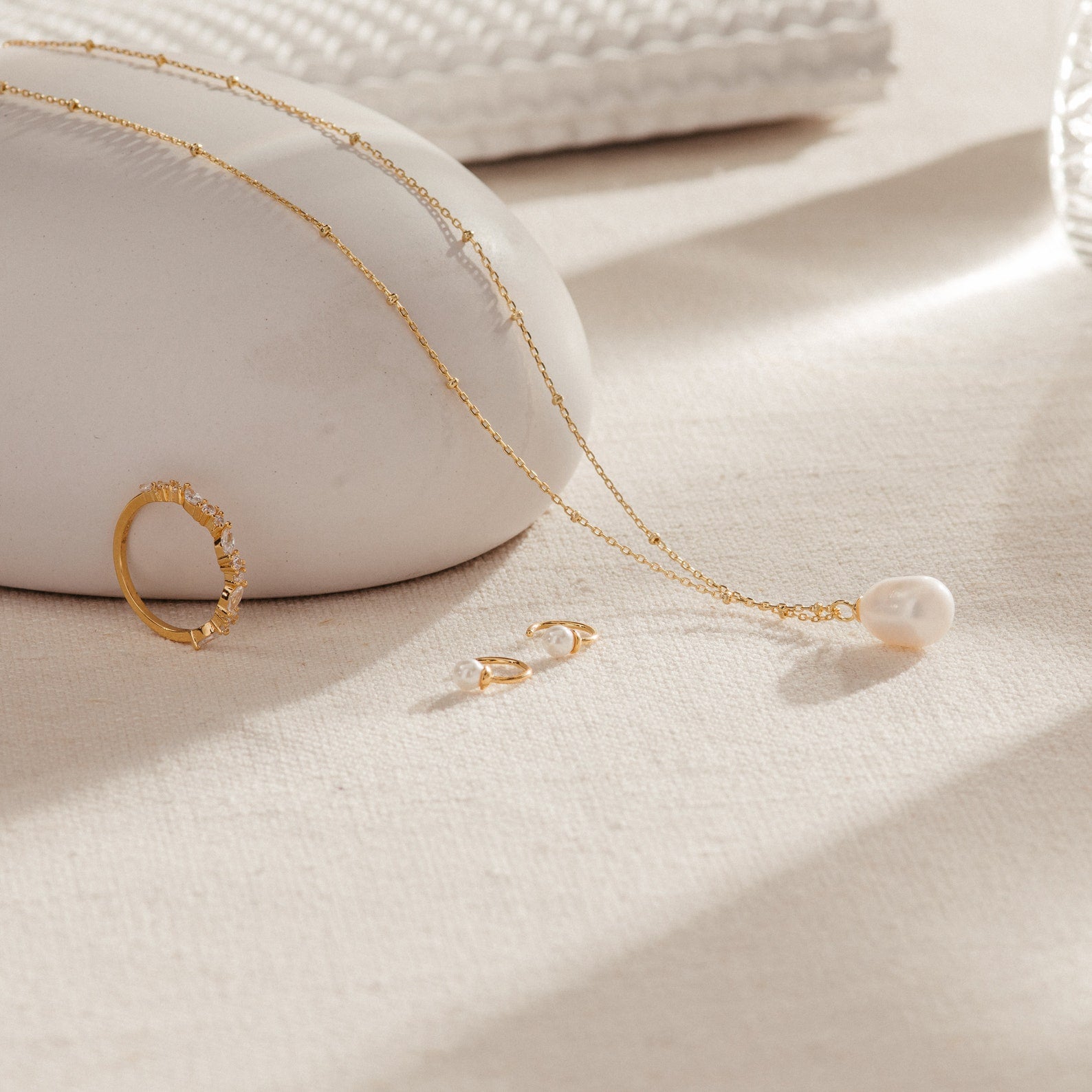 The Lila Pearl Huggies, a gold ring, and a pearl pendant necklace rest on a light beige surface with soft natural lighting—an elegant display of modern jewelry.