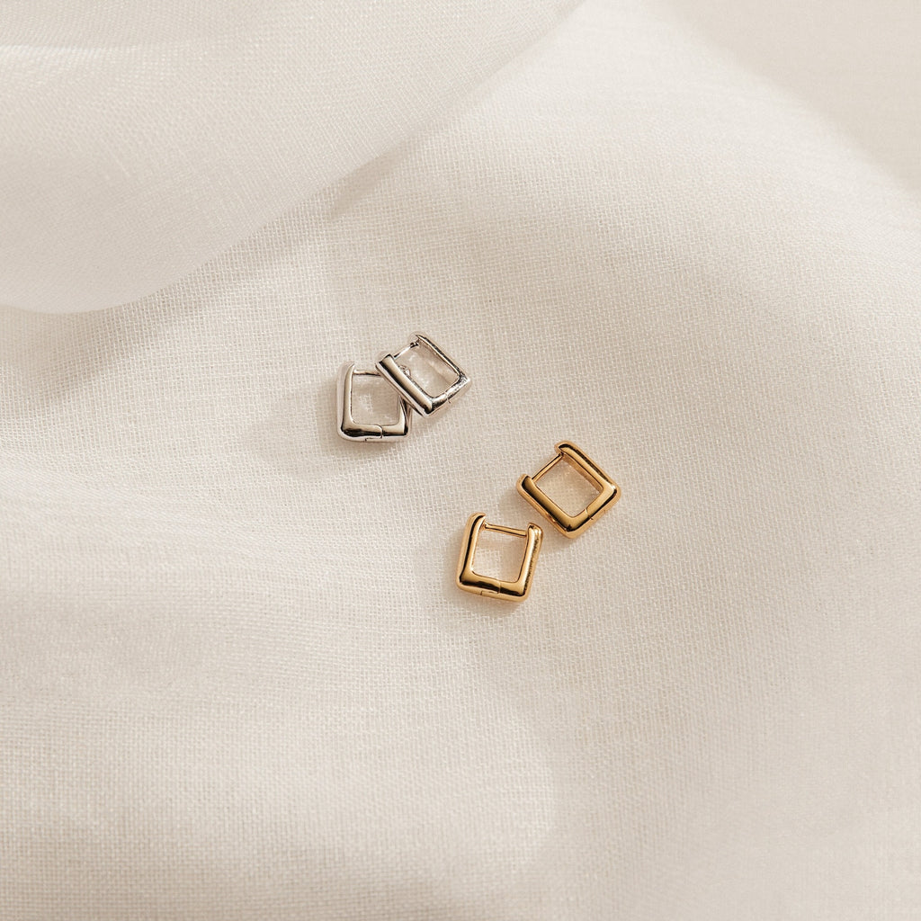 Two pairs of Kelsey Link Earrings, one silver and one gold, are displayed on soft white fabric. These contemporary hoop earrings feature clean geometric shapes for a modern look.