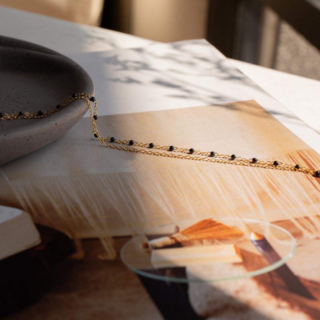 The Calypso Station Bracelet, a delicate gold chain with black beads ideal for stacking, is draped over a gray dish and magazine on a sunlit table.