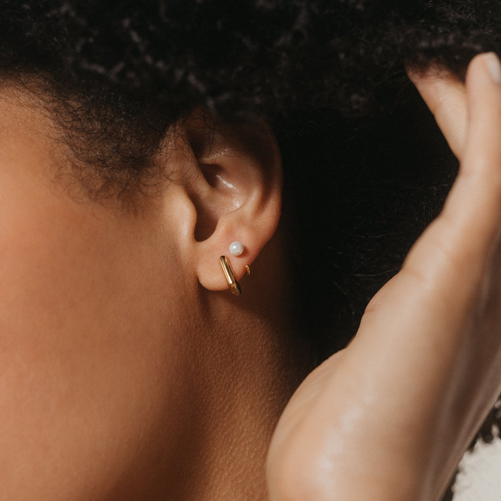 A close-up of an ear wearing the Lila Pearl Huggies—a dainty hoop with a small pearl accent—complemented by a hand softly touching the hair for a modern jewelry look.