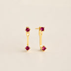 Straight‑on shot of a garnet pair gold birthstone ear jacket set—two small round CZ stones linked by a delicate bar—laid flat on a neutral background.