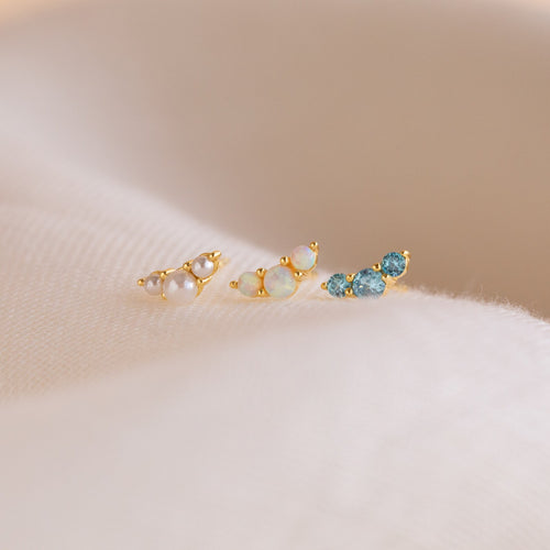 Three Bella Birthstone Studs—delicate gold earrings with pearls, opals, and blue gemstones—make thoughtful gifts for mothers and daughters, displayed on a soft cream fabric background.