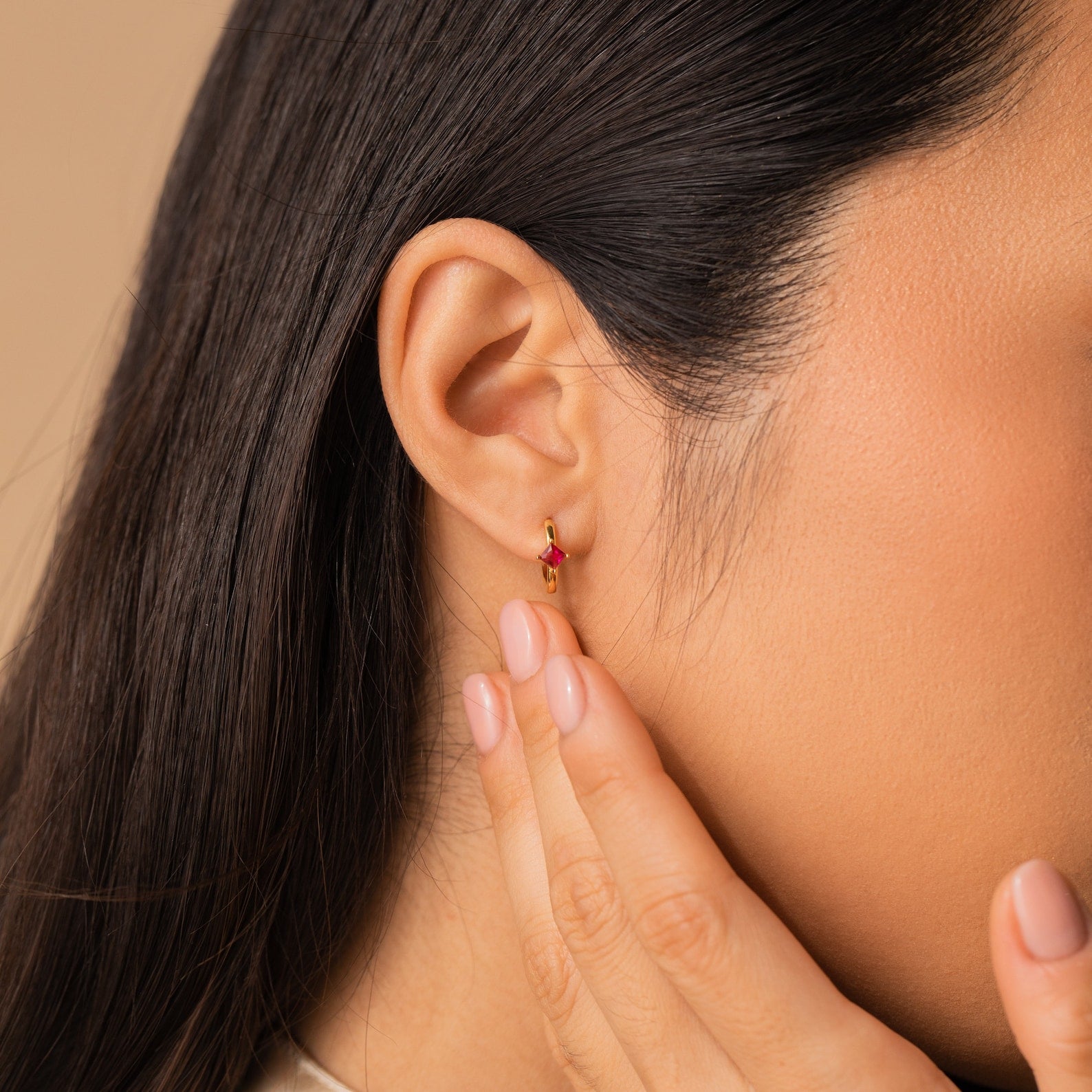 Close-up of a woman's ear wearing Athena Topaz Huggies, her hand touching her face and nails painted light pink.