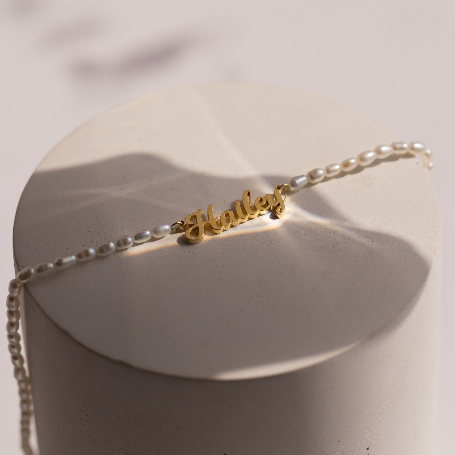 The Mellow Pearl Name Necklace features a gold dainty nameplate charm with pearl beads, elegantly displayed on a round, light-colored surface.