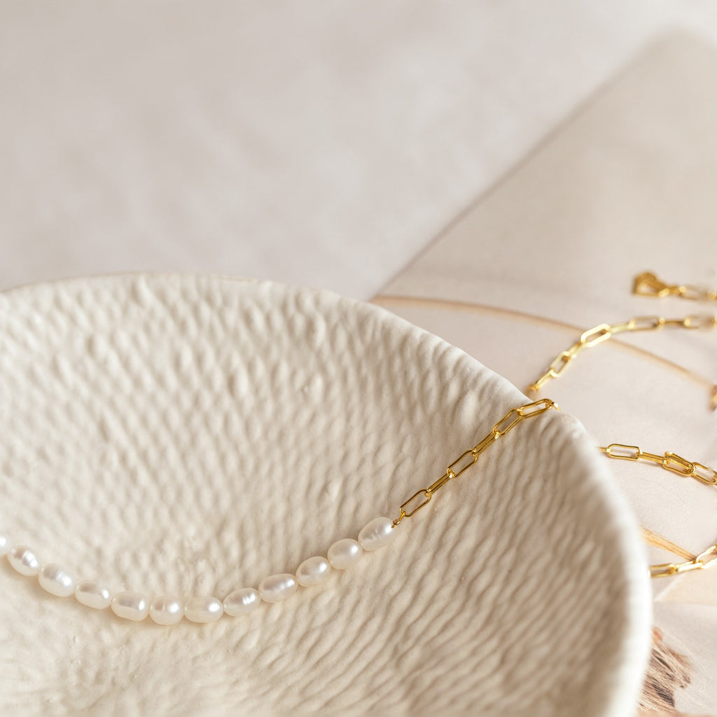 The Pearl & Paperclip Chain Necklace rests on a textured cream ceramic dish, its elegant dual design enhanced by the soft, neutral background.