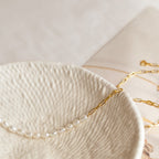 The Pearl & Paperclip Chain Necklace rests on a textured cream ceramic dish, its elegant dual design enhanced by the soft, neutral background.
