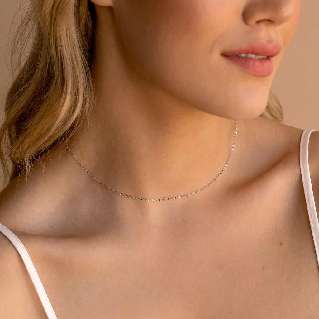 Woman wearing a delicate silver chain choker with small faceted links, styled with a white spaghetti strap top.