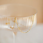 Delicate gold chain necklace with small faceted links draped over the rim of a clear ribbed glass coupe.
