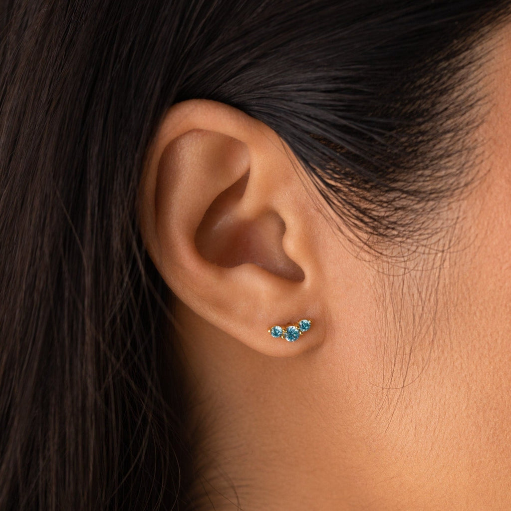 Close-up of a woman's ear wearing Bella Birthstone Studs—gold earrings with three small blue gemstones, ideal personalized jewelry for mothers and daughters.