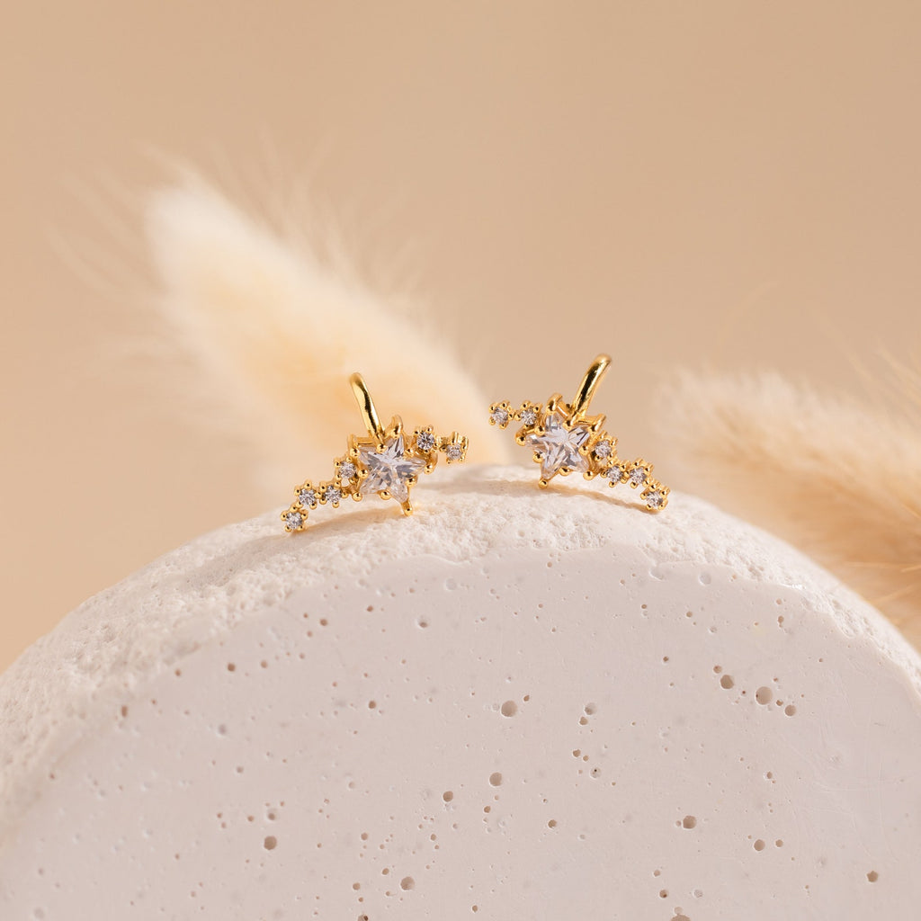 The Starry Night Ear Cuffs are gold, star-shaped cuffs with small crystals, elegantly displayed on a textured white stone and soft beige background. These no-piercing earrings add a sparkling touch to any look.