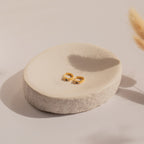 The Angelica Sapphire Huggies, gold rings set with blue gemstones, are displayed on a round, textured ceramic dish in soft natural light and paired elegantly with minimal earrings.