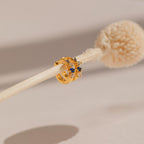 The Angelica Sapphire Huggies, featuring blue and clear gemstones in a minimalistic hoop design, are elegantly displayed on a dried flower stem against a neutral background.