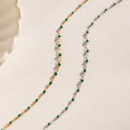 The Emerald Station Necklace, featuring delicate chains with emerald green beads in both gold and silver, adds beaded boho charm—beautifully displayed on a white surface.