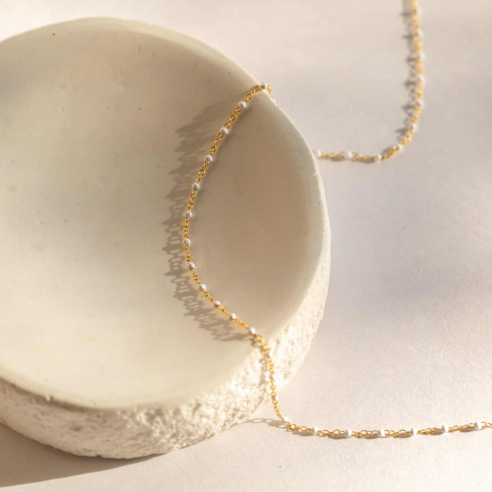 A Beaded Station Necklace draped over a textured, off-white ceramic bowl in soft sunlight, highlighting the playful elegance of this jewelry piece.