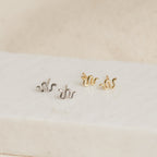The Tiny Python Studs, featuring one silver and one gold wavy pair, are displayed on a white rectangular surface—ideal for those who love dainty or boho-luxe jewelry.