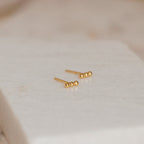Minimalist and perfect for daily wear, the Trio Beaded Bar Studs feature three polished gold beads, shown on a textured white background.