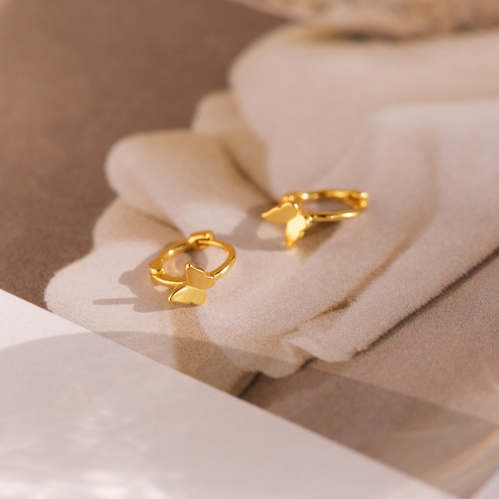 Flat lay of a pair of small gold butterfly cartilage huggies over a neutral surface highlighting small size and vibrant gold tones. 