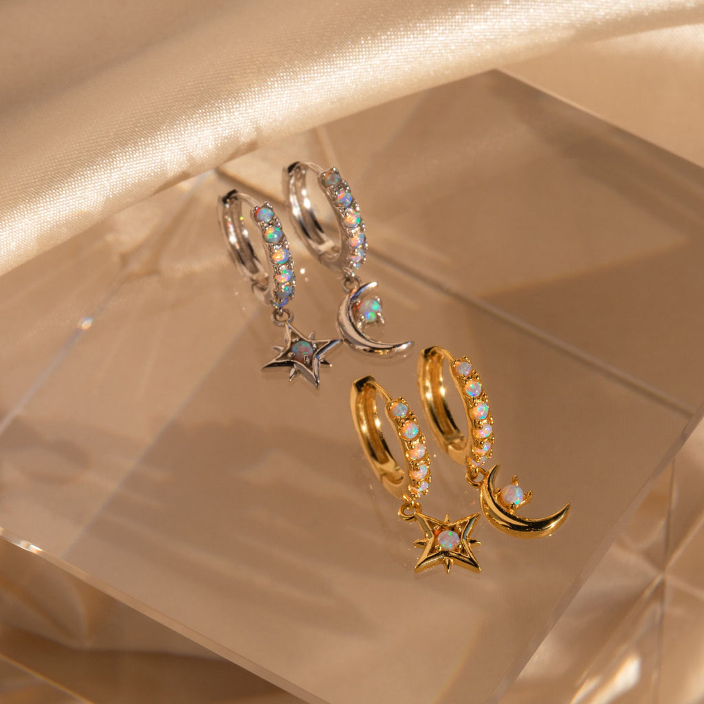 Two pairs of Astra Moon & Star Huggies—one silver, one gold—featuring celestial charms are displayed on a clear block, bringing lunar and starry style to your collection.