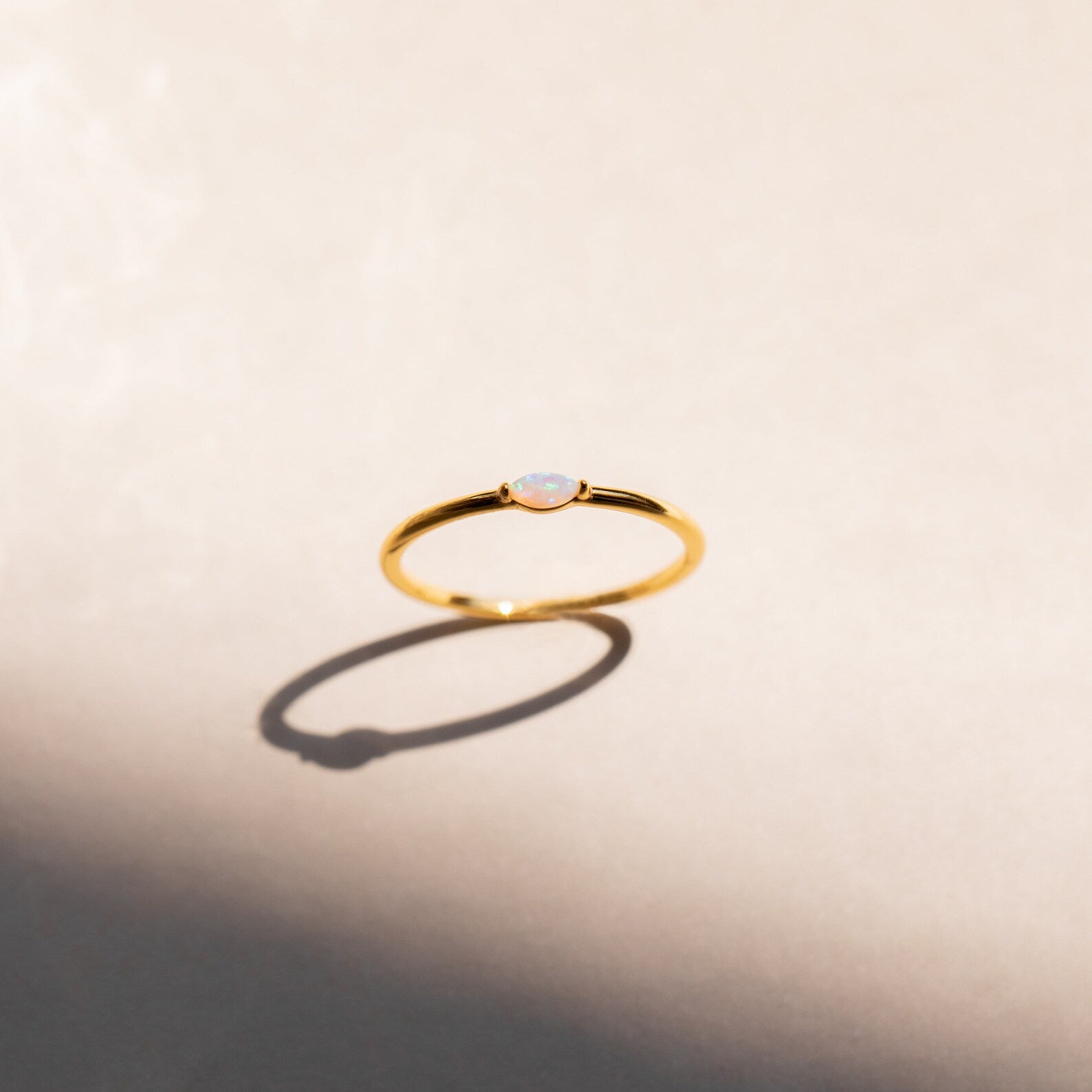 The Eloise Opal Ring features a delicate gold band and a small iridescent opal, casting subtle shadows on a light beige surface—a beautiful nod to minimalism.