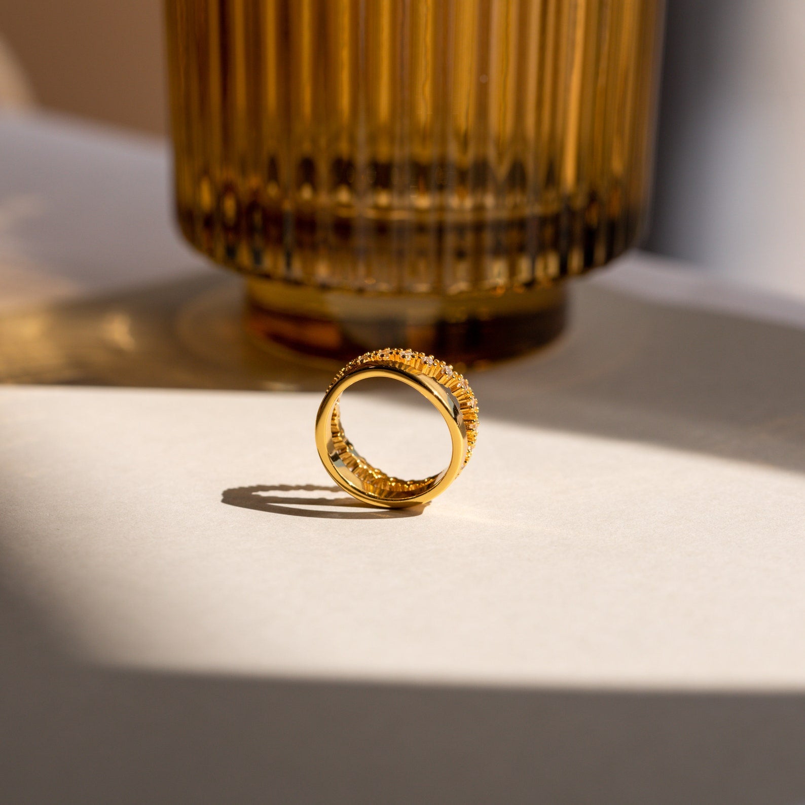The Wave Diamond Duo Ring stands upright on a sunlit surface near a ribbed amber glass vase.