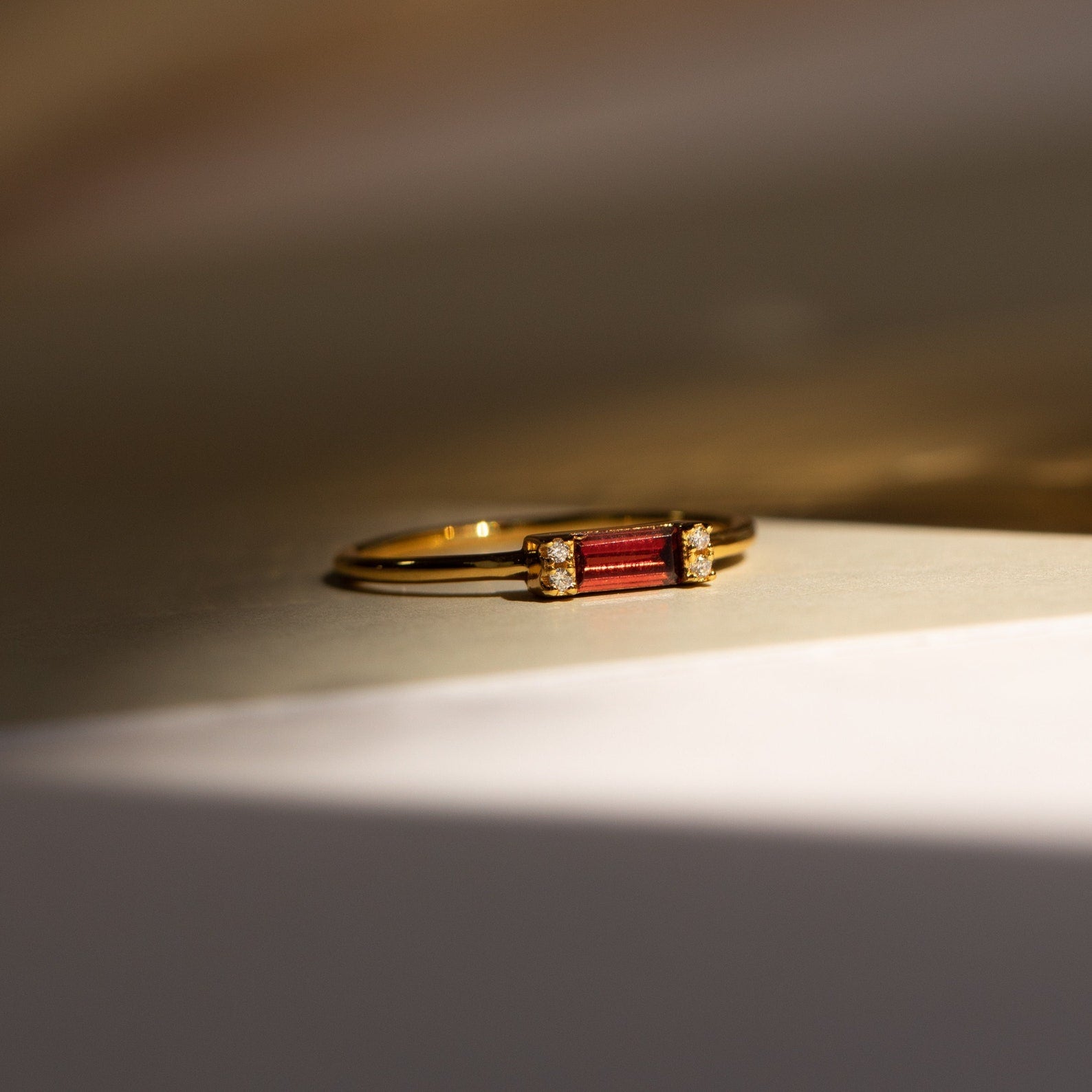 The Empire Garnet Ring features a gold band with a deep red rectangular baguette garnet and small diamonds, shown on a neutral surface in soft light.