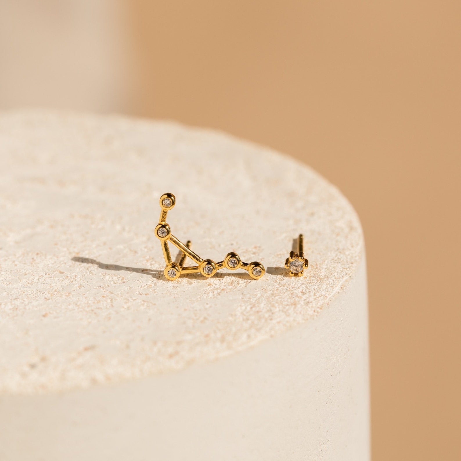 Constellation Earrings inspired by astrology, featuring gold constellation shapes with small gems, displayed on a textured cream-colored surface.