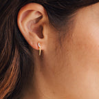 Close-up of a woman's ear wearing the Wren Spike Studs—small gold earrings with a clear CZ baguette diamond and a pointed spike drop charm in a lightweight design.