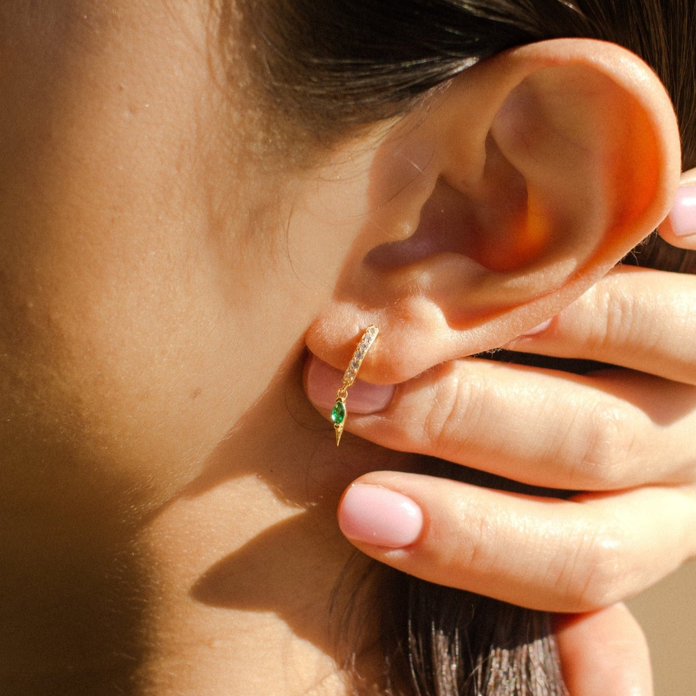 Close-up of a person wearing Raine Emerald Dagger Huggies with a green pendant, hand touching their ear—an ideal unique gift for her.