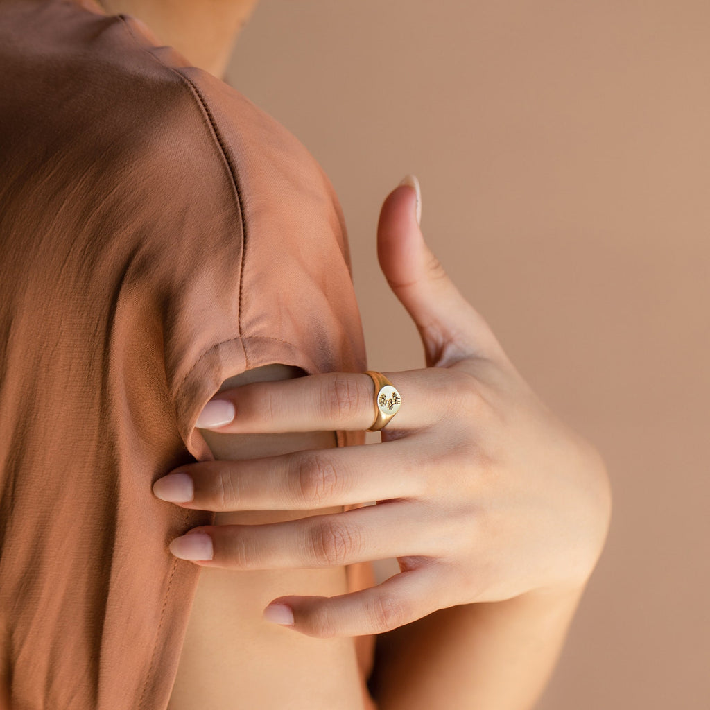 A person in a light brown top showcases the Birth Flower Signet Ring, featuring an engraved design in gold, on their finger.